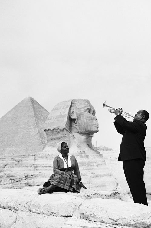 Louis Armstrong plays the trumpet while his wife sits listening with the Sphinx behind her, during a visit to the Pyramids at Giza in Egypt, 1961.